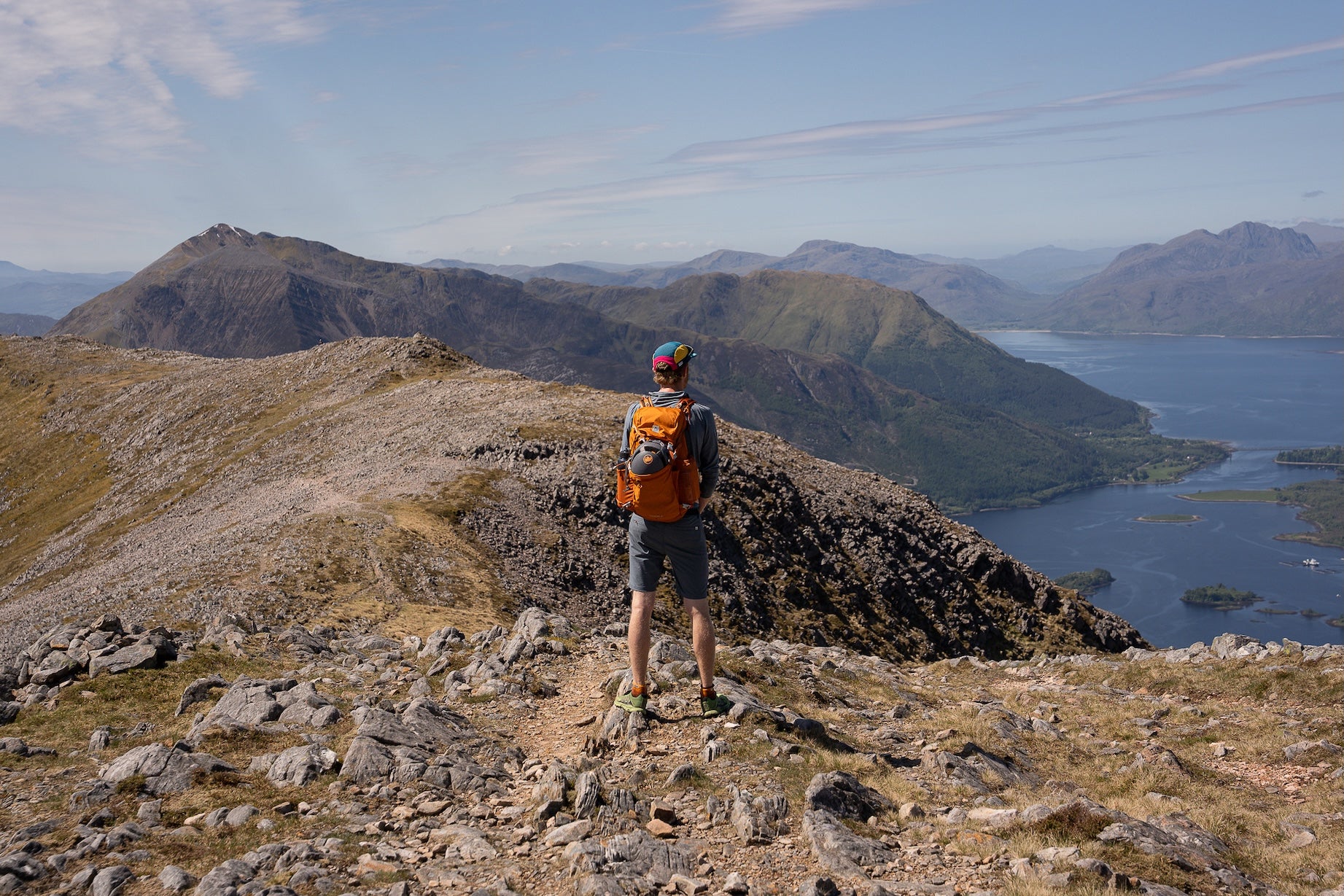Person with an orange backpack standing on a mountain peak overlooking a lake and mountains.